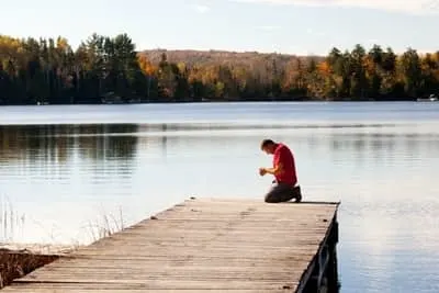 man kneeling on a dock near water, praying