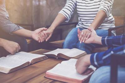 3 people around a coffee table holding hands praying with open bibles on the table.