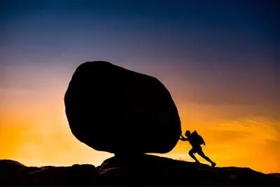 man pushing boulder while sun sets in the background